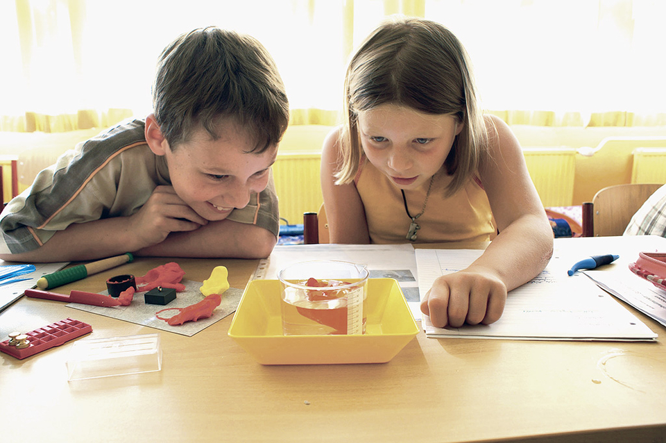 Das Bild zeigt zwei Kinder, die gespannt auf ein Experiment mit Wasser in einem transparenten Behälter blicken. Auf dem Tisch liegen verschiedene Bastelmaterialien und Unterlagen, die auf kreative Aktivitäten hinweisen.