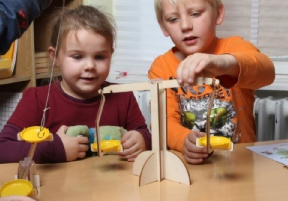 Das Bild zeigt zwei Kinder, die an einer Wippe experimentieren, um das Gleichgewicht zu prüfen. Sie halten jeweils eine gelbe Schaufel in der Hand, während sie gespannt beobachten, wie sich die Wippe bewegt.