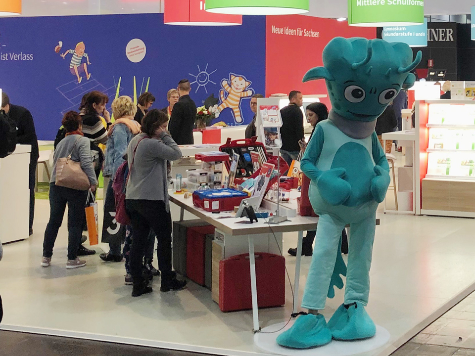 Das Bild zeigt einen Messestand für Experimentierbedarf mit mehreren Erwachsenen, die an einem Tisch interessiert Produkte betrachten. Im Vordergrund steht eine Person in einem grünen Kostüm, die das Interesse der Besucher auf sich zieht.