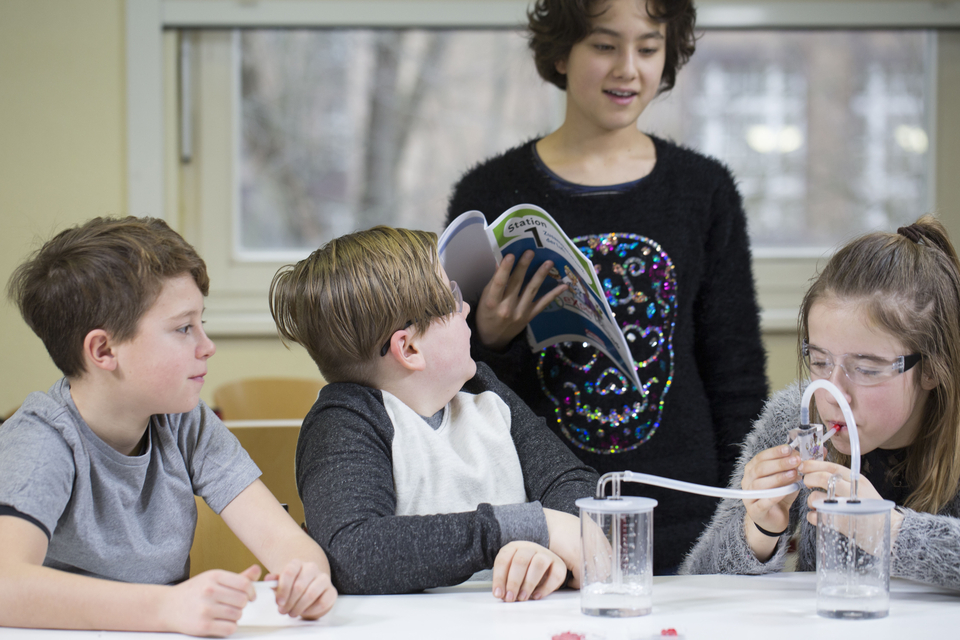 Das Bild zeigt eine Gruppe von Kindern, die an einem Experiment arbeiten. Ein Mädchen erklärt etwas aus einem Buch, während ein anderes Kind mit einem wissenschaftlichen Gerät in zwei Bechern experimentiert.