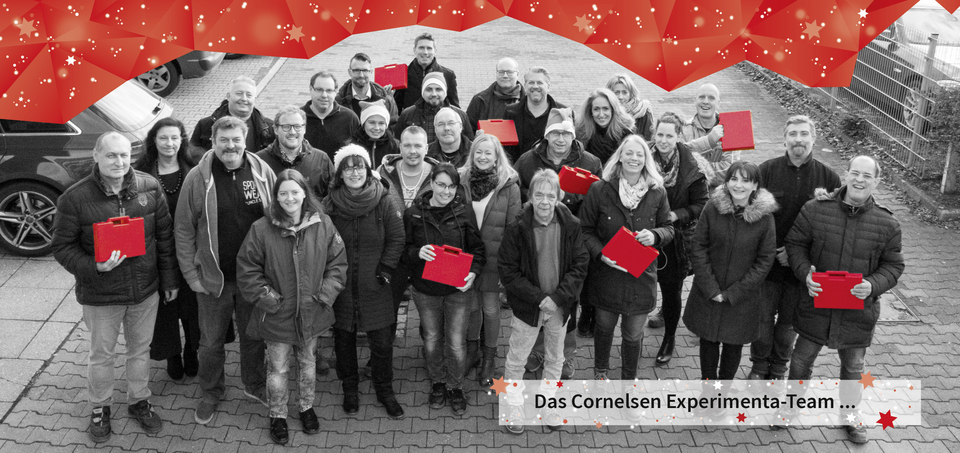 Das Bild zeigt das Cornelsen Experimenta-Team, welches in einer Gruppe auf einem Parkplatz steht. Die Mitarbeiterinnen und Mitarbeiter halten rote Koffer in den Händen und sind in einem informellen, freundlichen Rahmen zusammengekommen.