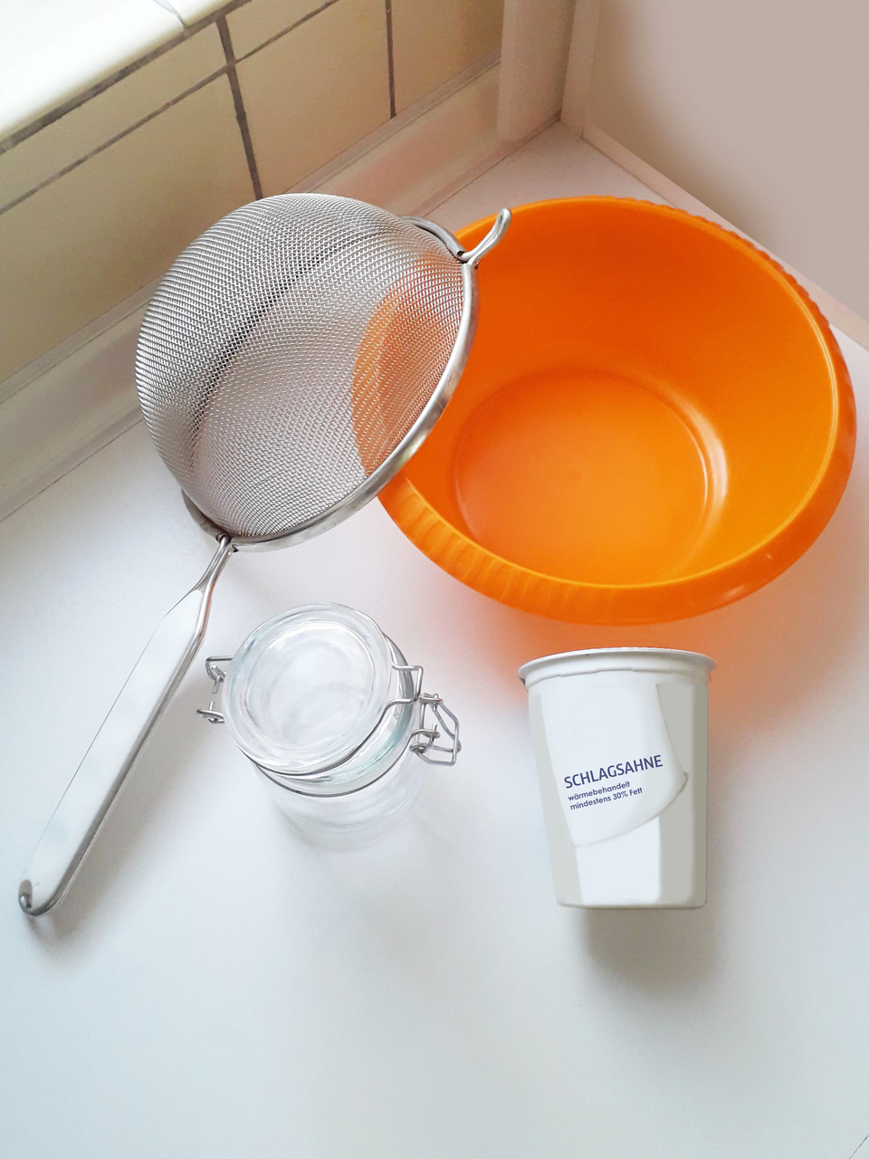 Das Bild zeigt eine orangefarbene Schüssel, ein feinmaschiges Sieb, ein rundes Glasgefäß mit Verschluss und einen Becher mit der Aufschrift „Schlagsahne“. Dies sind Materialien und Bestandteile, um Butter herzustellen.