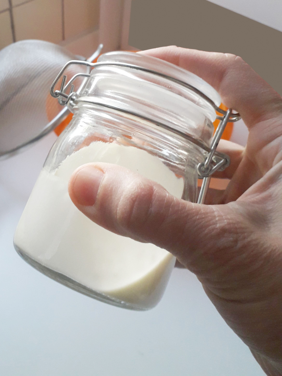 Das Bild zeigt eine Hand, die ein Glas mit Sahne hält. Im Hintergrund ist ein Sieb zu sehen, das auf ein Experiment hindeutet wie die Herstellung von Buttermilch oder Butter.