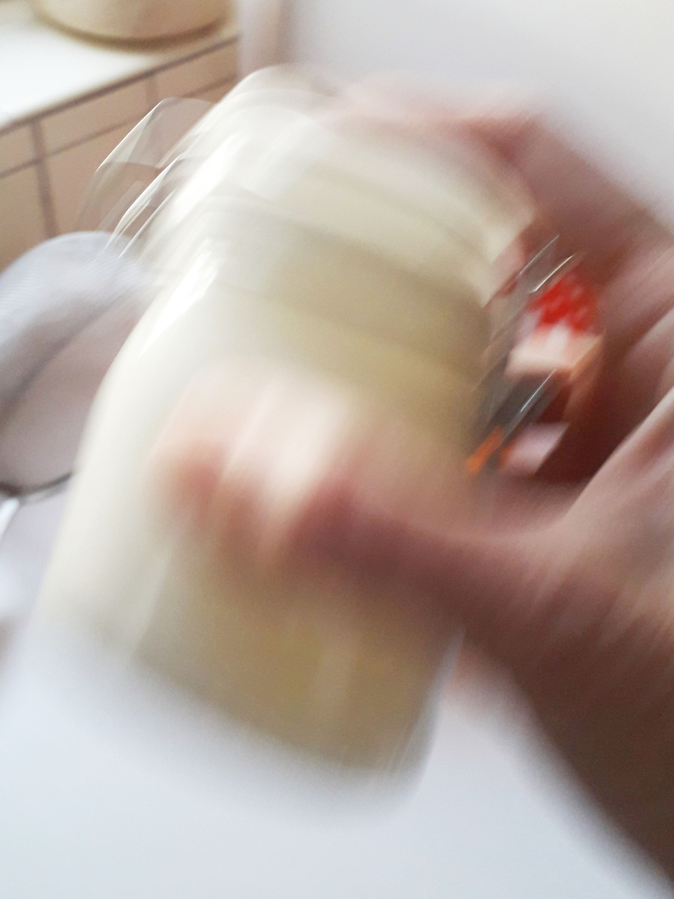 Das Bild zeigt eine verschwommene Hand, die ein Glas hält, während es geschüttelt wird. Im Hintergrund sind unscharfe Küchenelemente zu erkennen, was auf eine Experimentier- oder Kochsituation hindeutet.