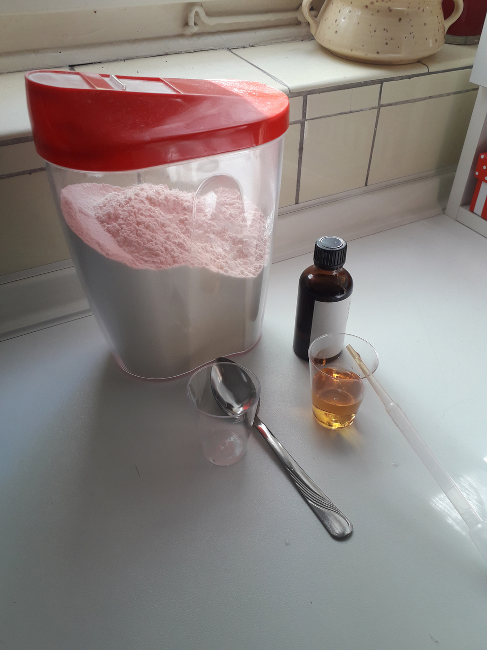 Das Bild zeigt einen großen Behälter mit pinkem Pulver, daneben ein braunes Fläschchen und zwei Messbecher, einer gebogen und einer transparent mit einer gelben Flüssigkeit. Eine Esslöffel liegt ebenfalls auf dem Tisch, bereit für Experimente.