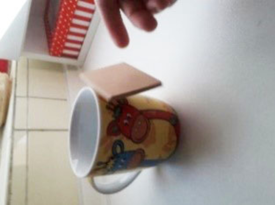 Das Bild zeigt eine bunte Tasse mit Tiermotiven, die auf einem Tisch steht. An der Tasse rechts befindet sich ein kleines Holzstück, das zu schweben scheint, daneben ist noch eine Hand zu erkennen, die das Holzstück nicht festhält.