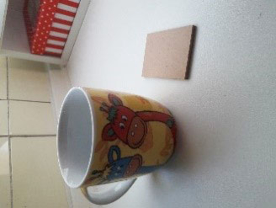 Auf dem Bild ist eine bunte Tasse mit Kuh-Motiven und ein rechteckiges Stück Pappe zu sehen. Der Hintergrund zeigt eine helle Oberfläche, die in einem pädagogischen Kontext genutzt werden könnte.