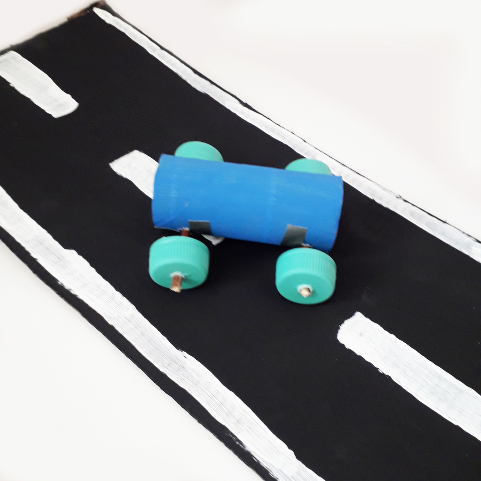 Das Bild zeigt ein selbstgebautes Fahrzeug aus recycelten Materialien, das auf einer kunstvoll gestalteten Straße platziert ist. Das Fahrzeug besteht aus einem blauen Zylinder mit grünen Radachsen, während die Straße schwarz mit weißen Linien dargestellt 