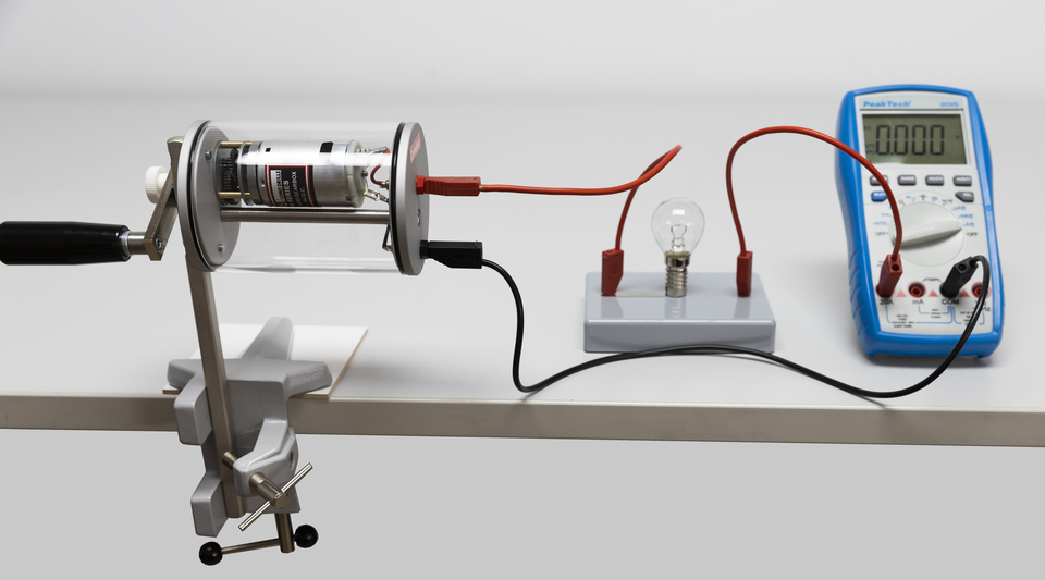 Das Bild zeigt ein Experimentiergerät, den Dynamot von Cornelsen Experimenta, mit einem Motor, der mit einer Glühbirne und einem Multimeter verbunden ist. Die Anordnung dient zur Veranschaulichung elektrischer Stromkreise und der Messung von Stromstärke.