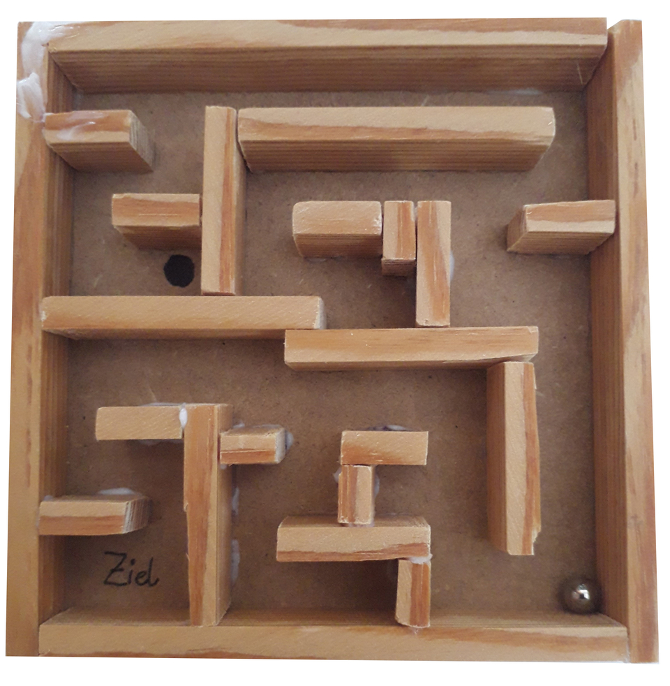 Das Bild zeigt ein einfaches selbstgebautes Labyrinth aus Holz mit verschiedenen Stäben, durch die eine kleine Metallkugel hindurchrollen kann. Am unteren Ende ist ein markierter Zielpunkt zu erkennen, wo die Kugel ankommen soll.