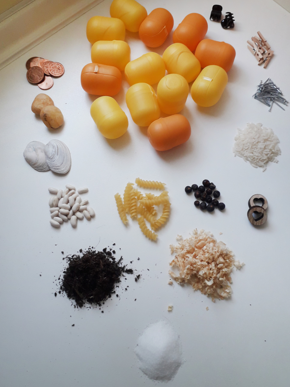 Das Bild zeigt eine Vielzahl von Materialien, die verwendet werden können, um ein Hörmemory zu bauen, darunter kleine bunte gelbe Plastikbehälter, verschiedene Lebensmittel wie Reis und Pasta, sowie Gegenstände wie Münzen und Nägel. Diese Materialien förd