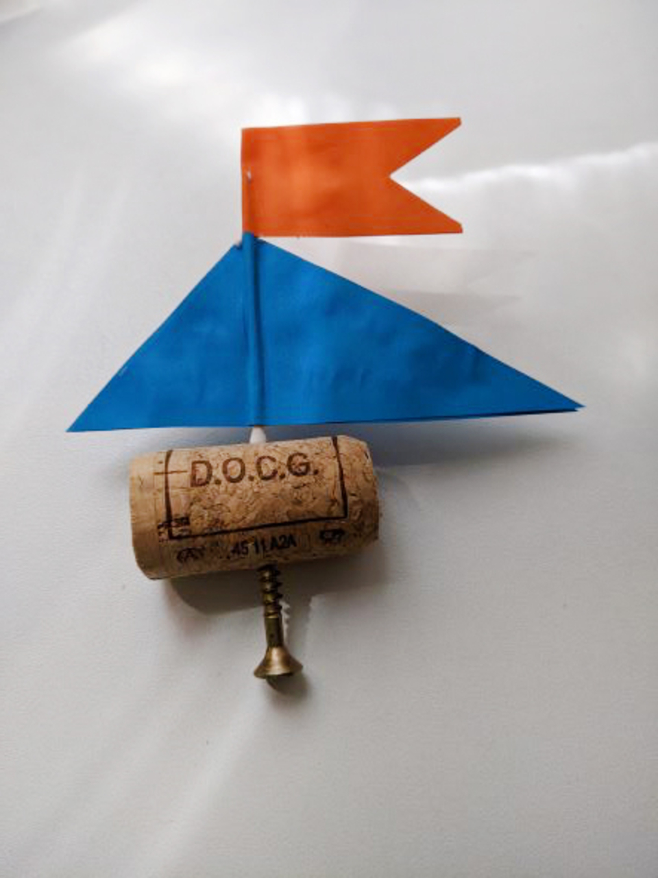 Das Bild zeigt ein einfaches Bootmodell, das aus einem Korken als Rumpf und bunten Papierdreiecken als Segeln besteht. Die blaue und orangefarbene Flagge verleihen dem Modell einen ansprechenden Look für experimentelle Zwecke im Bildungsbereich.