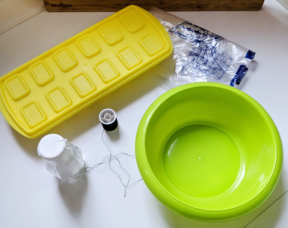 Das Bild zeigt eine Sammlung von Experimentiermaterialien, darunter eine grüne Schüssel, eine gelbe Silikonform, eine kleine Glasflasche und einen Garnrollen. Diese Gegenstände sind für verschiedene Bildungs- und Experimentieraktivitäten gedacht.
