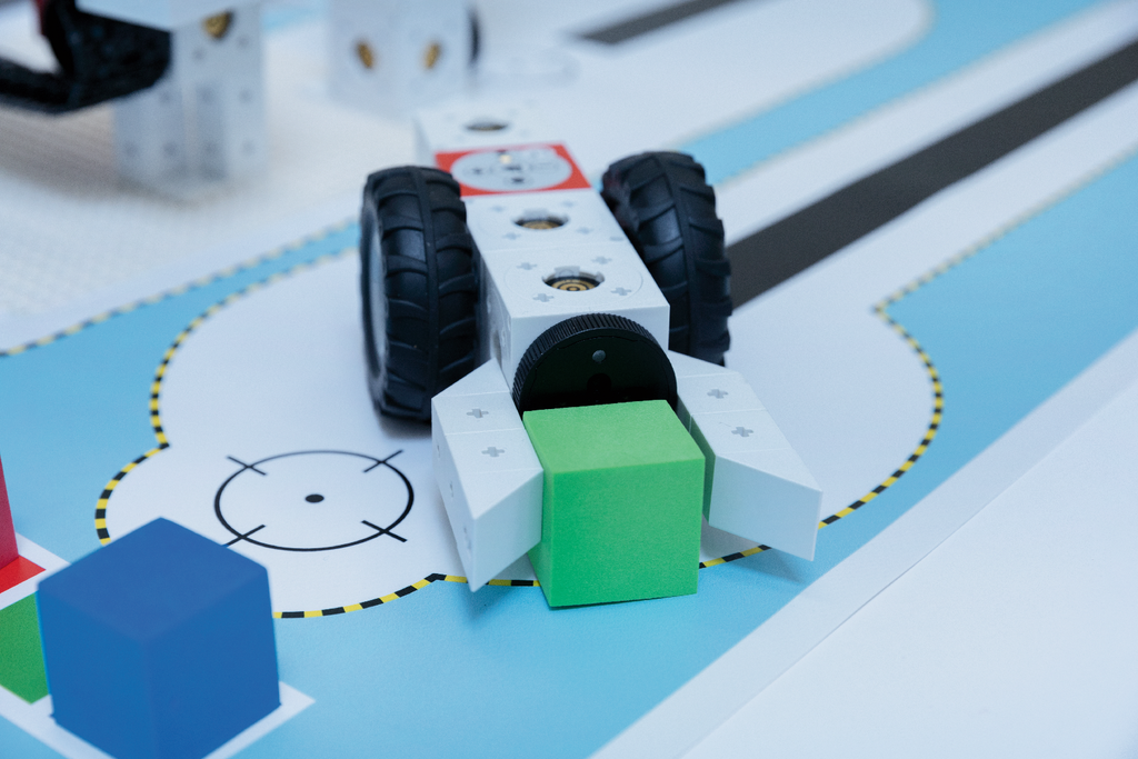 Das Bild zeigt einen Roboter mit großen Reifen, der offenbar einen grünen Würfel transportiert. Im Hintergrund ist eine farbige, mit verschiedenen Formen und einer Straße gestaltete Experimentierfläche zu sehen.