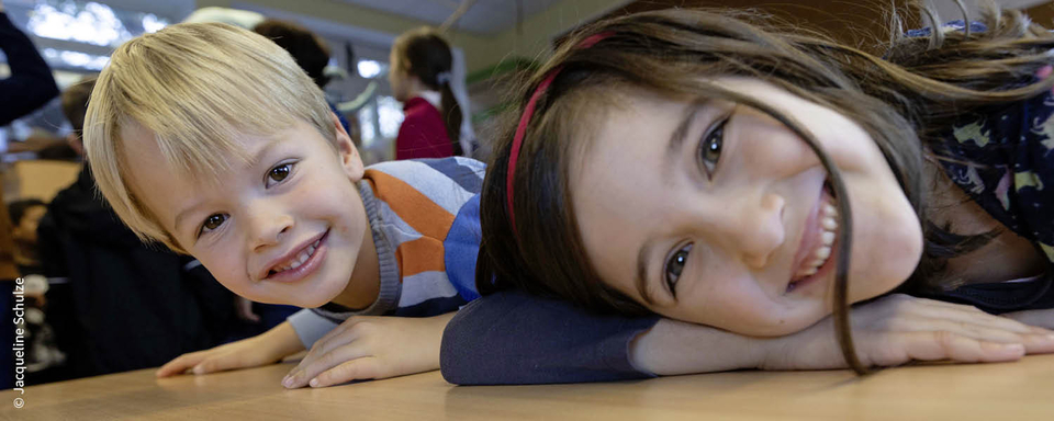 Das Bild zeigt zwei fr&ouml;hliche Kinder, einen Jungen und ein M&auml;dchen, die eng beieinander ihre Gesichter auf einem Tisch ablegen und in die Kamera l&auml;cheln. Im Hintergrund sind weitere Kinder und eine helle, freundliche Umgebung zu sehen.