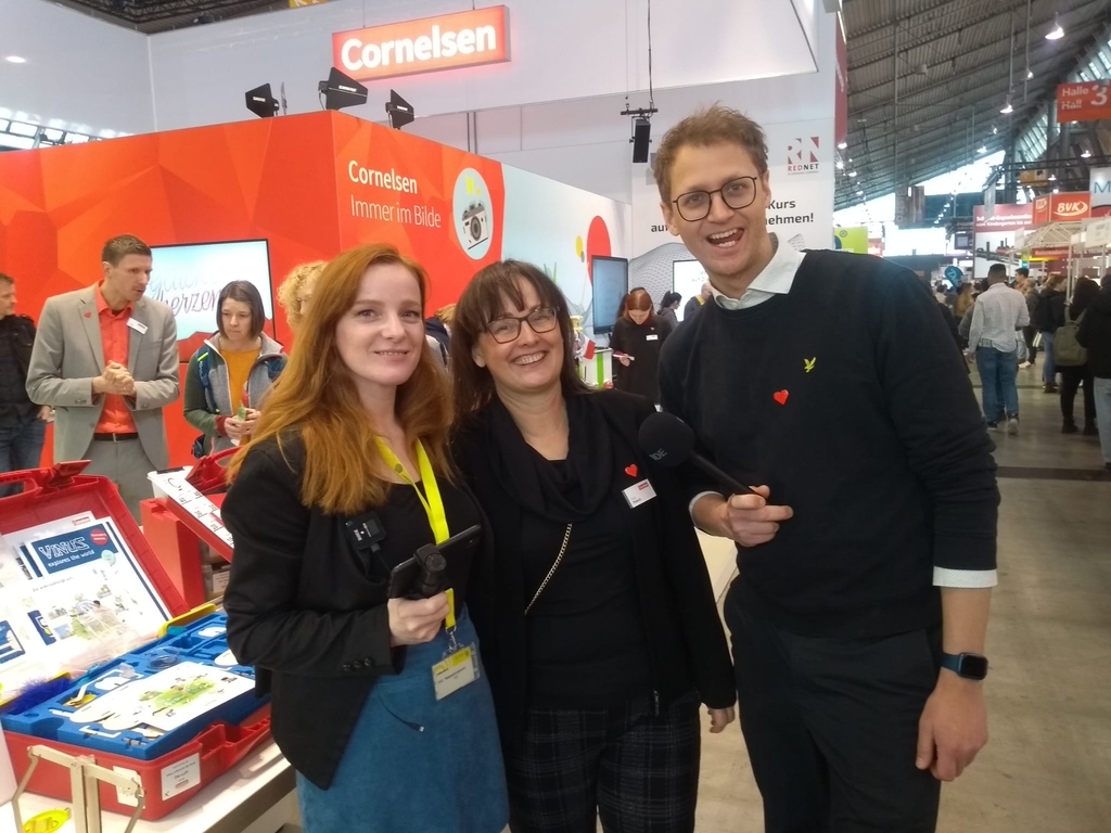 Das Bild zeigt mehrere Personen an einem Messestand von Cornelsen, der sich mit Experimentierbedarf für das Bildungswesen beschäftigt. Im Vordergrund lächeln zwei Frauen und ein Mann in formeller Kleidung in die Kamera, während im Hintergrund weitere Besucher zu sehen sind.