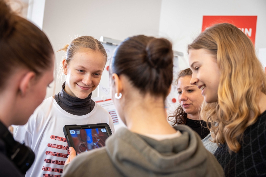 Auf dem Bild sind mehrere Mädchen zu sehen, die fasziniert auf ein Tablet schauen, das mithilfe von Augmented Reality das Körperinnere zeigt. Ein Mädchen hat das Magic T-Shirt an und lächelt, während die anderen aufmerksam dem Inhalt auf dem Bildschirm fo