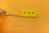 Das Bild zeigt ein gelbes Experimentiergerät mit einer flexiblen Metallspitze, das zur Durchführung von physikalischen Experimenten verwendet wird. Daneben liegt ein schlanker Metallstab, der möglicherweise als zusätzlicher Bestandteil für das Experiment 