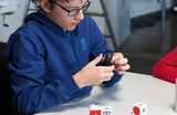 Ein Junge mit Brille untersucht ein kleines Gummirad, während er an einem Tisch sitzt. Vor ihm liegen verschiedene Bauklötze in Weiß und Rot, die möglicherweise für ein Experiment oder ein Projekt im Bildungsbereich gedacht sind.