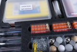 Das Bild zeigt eine sortierte Experimentierbox mit verschiedenen Materialien, darunter Knete, Messzylinder, Gewichte und diverse Bauteile wie Kugeln und Holzstücke. Die Inhalte sind übersichtlich in Fächern angeordnet, ideal für schulische Experimente.