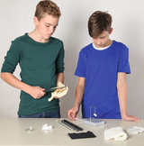 Das Bild zeigt zwei Jugendliche, die an einem Tisch mit verschiedenen Experimentiermaterialien arbeiten. Einer der Jungen hält ein Instrument, während der andere auf ein physikalisches Experiment mit einer Federwaage fokussiert ist.
