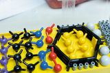 Das Bild zeigt eine bunte Modellbausatz-Anordnung von Molekülen und Atomen auf einer gelben Basis. Verschiedene Farben repräsentieren verschiedene chemische Elemente, was für den Experimentierbedarf im Bildungswesen geeignet ist.