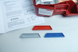 Das Bild zeigt eine Experimentierbox zum Thema „Sicherer Umgang mit Feuer“ mit farbigen Plättchen, die die Begriffe „Temperatur“, „Brennstoff“ und „Luft“ darstellen. Ebenso ist ein Handbuch mit Anleitungen für Experimente in der Box zu sehen.