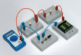 Das Bild zeigt eine elektrische Schaltung mit mehreren Komponenten, darunter Glühbirnen, Batterien und ein Multimeter. Die Kabel sind farblich kodiert und verbinden die einzelnen Bauteile zur Durchführung von Experimenten im Bereich Elektrotechnik.