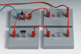 Das Bild zeigt ein Experimentier-Set mit einem Transistor und mehreren Kabeln in Rot und Blau, die an die Anschlüsse E, B und C angeschlossen sind. Es ist auf einer grauen Platte angeordnet, die für Bildungszwecke im Bereich Elektrotechnik verwendet wird.