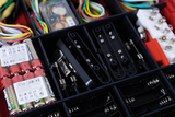 Das Bild zeigt eine Übersicht von Experimentiermaterialien in einem Kasten, der Batterien, Anschlussklemmen und verschiedene elektrische Bauteile enthält. Die farbigen Drähte und weitere Ergänzungen deuten auf ein breites Anwendungsspektrum im Bildungsbereich hin.