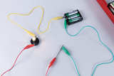 Das Bild zeigt eine elektrische Schaltung mit einer Glühlampe, die an eine Batterie angeschlossen ist. Bunte Kabel verbinden die Komponenten und ermöglichen einen praktischen Versuch zur Elektrizität im Bildungsbereich.
