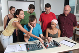 Auf dem Bild arbeiten mehrere Jugendliche und ein Lehrer gemeinsam an Experimentiermaterialien an einem Tisch. Sie scheinen sich auf eine technische Aufgabe zu konzentrieren, während sie miteinander diskutieren und Anweisungen austauschen.