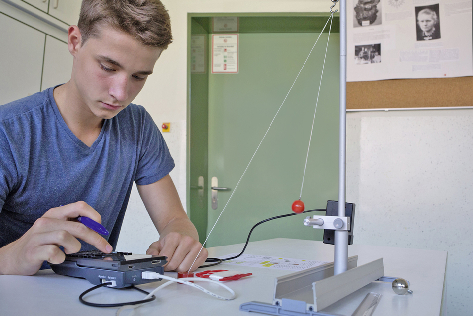 Ein Schüler arbeitet an einem Experiment, das mit einem physikalischen Versuchsaufbau zur Untersuchung von Kräften und Bewegungen zu tun hat. Neben ihm liegt ein Gerät, vermutlich zur Datenerfassung, während eine rote Kugel an einem Seil schwingt.