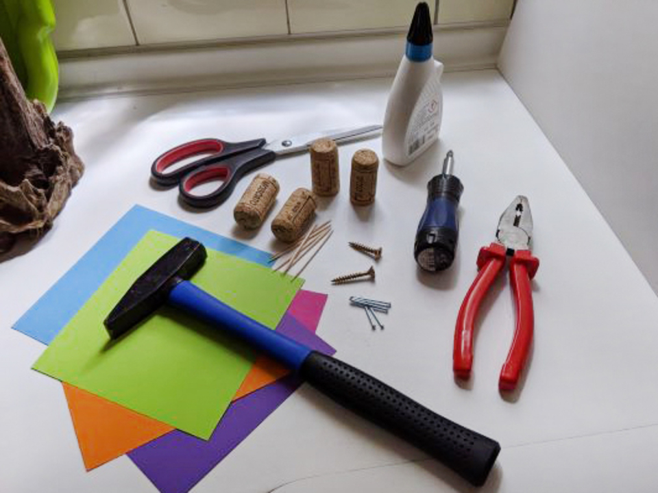 Vielfältige Materialien zum Experimentieren sind angeordnet, darunter bunte Bastelpapiere, eine Gummihammer, Scheren, Korken, Schrauben und verschiedene Werkzeuge. Diese Utensilien sind ideal für kreative Projekte im Bildungsbereich.
