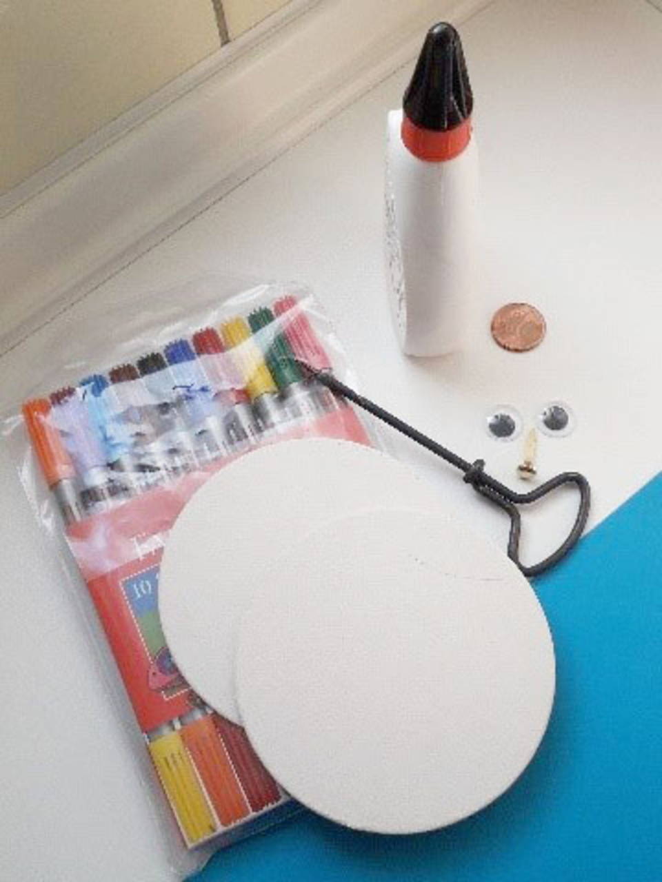 Im Vordergrund sind zwei runde, weiße Pappen mit einer Schlaufe, die einen Verbindungspunkt bietet. Daneben befinden sich eine Packung bunter Marker, eine Flasche Kleber, ein centstück und zwei Augen, die für kreatives Basteln verwendet werden können.