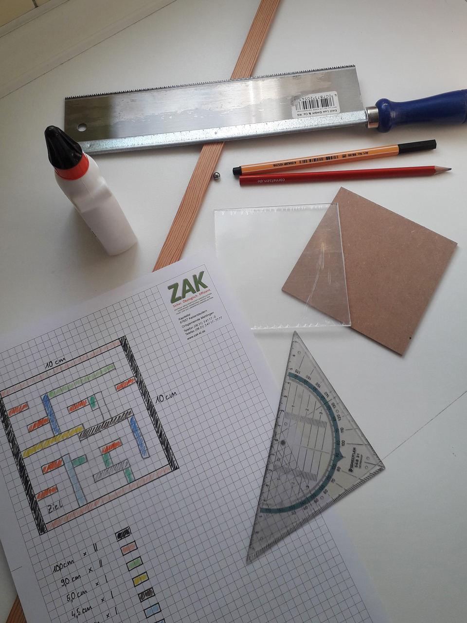 Auf dem Tisch liegen verschiedene Werkzeuge und Materialien, darunter eine Handsäge, ein Lineal, Kleber, Bleistifte, ein Geodreieck und Zeichenmaterialien. Zudem befindet sich eine Planungsskizze auf kariertem Papier, die eine Bauanleitung zeigt.