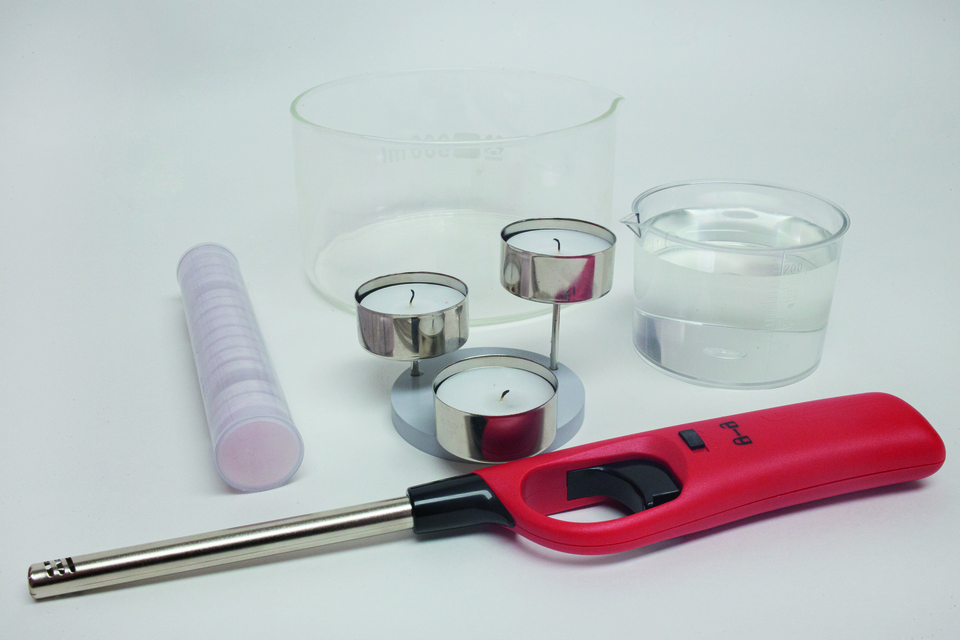 Im Vordergrund befinden sich ein roter Feuerzeug, drei Teelichter und eine transparente Wasserbecher. Daneben liegt ein röhrenförmiger Behälter und ein weiterer Messbecher mit klarem Wasser, die für Experimente im Bildungsbereich genutzt werden können.