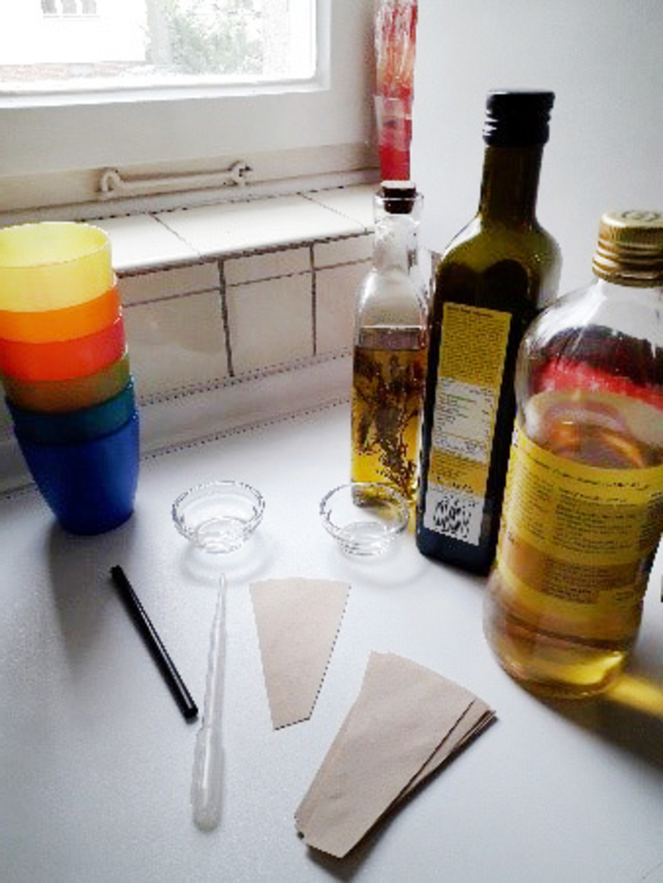 Farbenfrohe Becher stehen neben Flaschen mit verschiedenen Flüssigkeiten, darunter Olivenöl. Auf dem Tisch liegen zwei Papierstücke, eine Pipette und zwei kleine Schalen, die auf ein Experiment hinweisen.