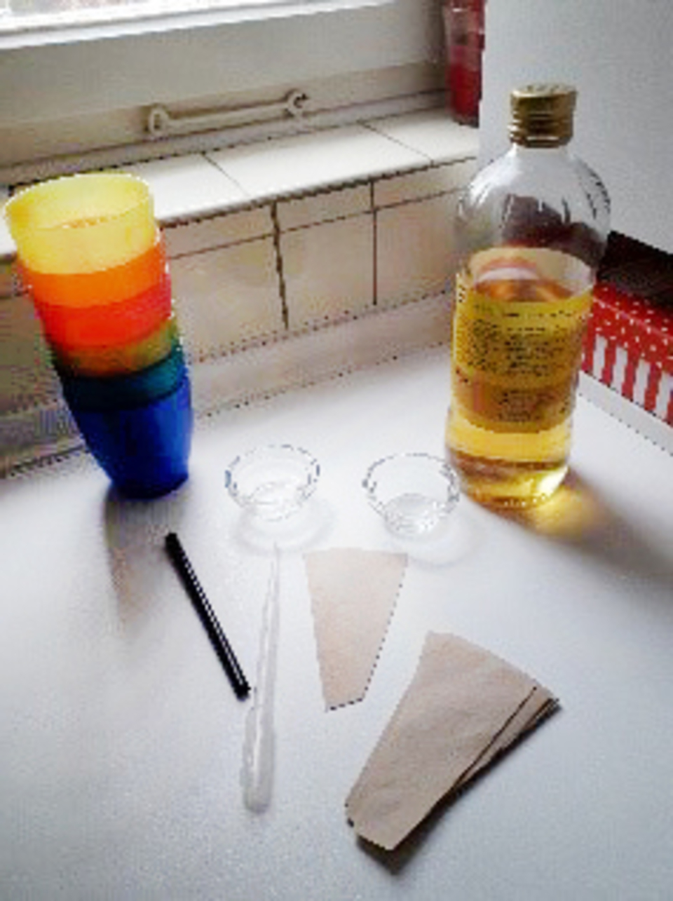 Bunte Becher aus Kunststoff stehen gestapelt neben einer Flasche mit Flüssigkeit und zwei kleinen Schalen. Außerdem sind ein Strohhalm und zwei Papiertüten auf dem Tisch verteilt, die für Experimente im Bildungsbereich verwendet werden können.