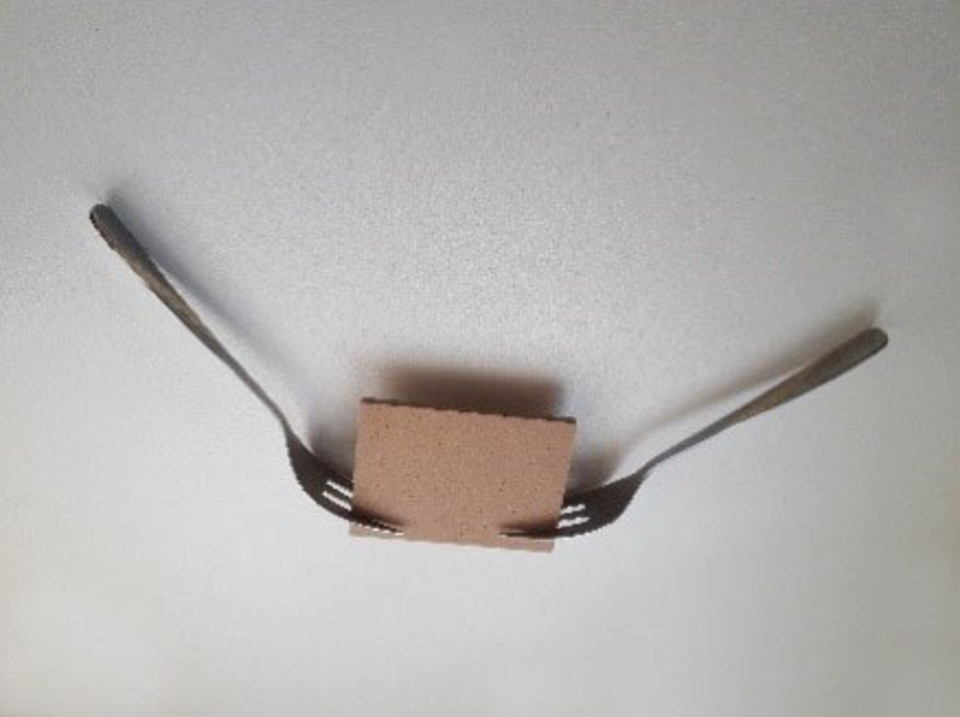 Zwei Gabeln sind an einem kleinen, quadratischen Stück Karton befestigt, das in der Mitte des Bildes liegt. Die Gabeln ragen nach außen und scheinen eine Art Halterung oder Unterstützung für das Kartonstück zu bilden.