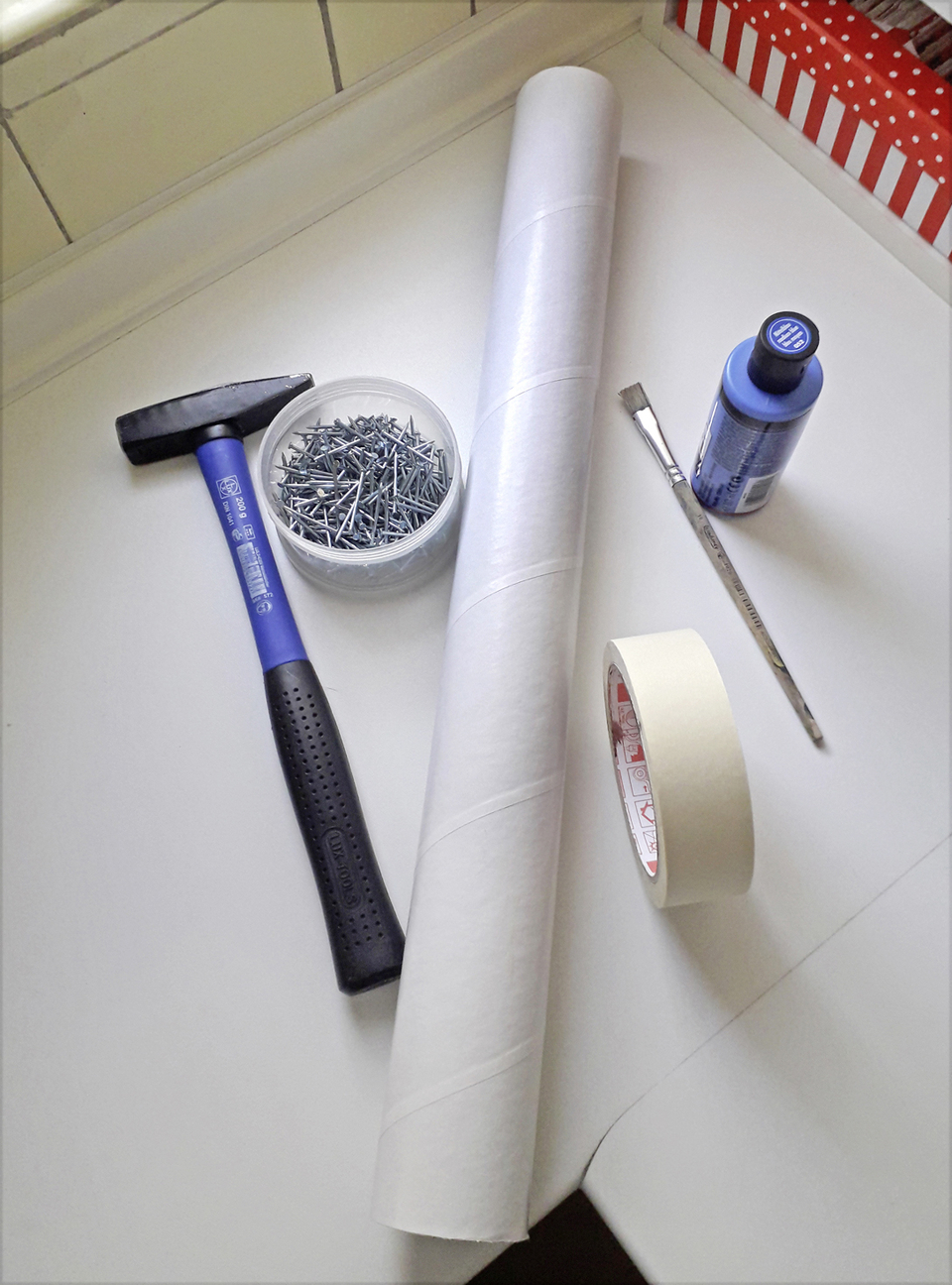 Auf der Arbeitsfläche sind verschiedene Materialien für experimentelle Projekte angeordnet. Zu sehen sind ein Hammer, eine Schachtel mit Nägeln, ein Rolle Papier, Buntfarbe samt Pinsel sowie Klebeband, alles für kreative Aktivitäten im Bildungsbereich.