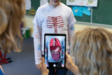 Eine Schülerin trägt ein T-Shirt, das das menschliche Skelett zeigt, während eine Lehrkraft mit einemTablet die inneren Organe in einer Augmented-Reality-Anwendung anzeigt. Dies dient als interaktive Lernmethode zur Veranschaulichung anatomischer Zusammenhänge.