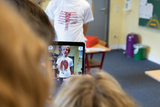 Ein Schüler präsentiert ein T-Shirt mit einem bildlichen Diagramm des menschlichen Körpers, während ein anderer Schüler ihn mit einem Tablet aufnimmt. Im Hintergrund sind Klassenräume und Möbel zu sehen, die eine aktive Lernsituation darstellen.