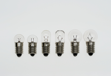 Sechs verschiedene Glühbirnen mit Schraubsockel sind auf einer weißen Fläche angeordnet. Die Birnen variieren in Größe und Form und zeigen die unterschiedlichen inneren Strukturen, die für ihre Funktion als Lichtquelle verantwortlich sind.