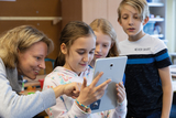 Eine Lehrerin zeigt einer Gruppe von Kindern ein Tablet, auf dem sie aufmerksam etwas betrachten. Die Kinder wirken interessiert und engagiert, während sie miteinander und mit der Lehrerin interagieren.