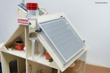 Ein Experimentieraufbau mit einem durchsichtigen Behälter und einer roten Kappe steht im Vordergrund. Im Hintergrund befindet sich ein Solarpanel, das auf einem hölzernen Modell mit verschiedenen Elementen, darunter Pflanzen und ein Thermometer, montiert ist.