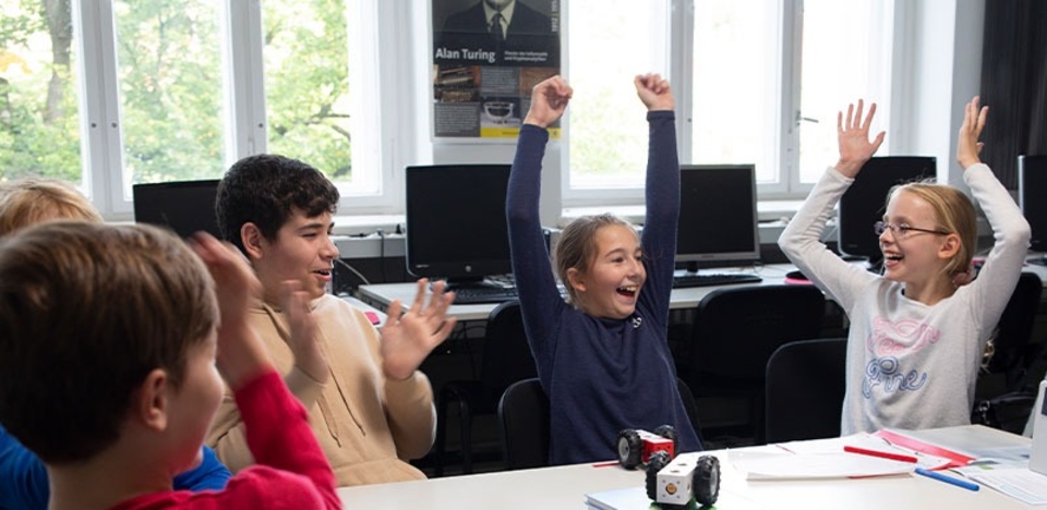 Eine Gruppe von Kindern sitzt an einem Tisch und freut sich sichtbar &uuml;ber ein Experiment oder eine erfolgreiche Aktivit&auml;t. Ihre positiven Reaktionen, wie Jubel und lachende Gesichter, spiegeln Begeisterung f&uuml;r das Lernen wider.