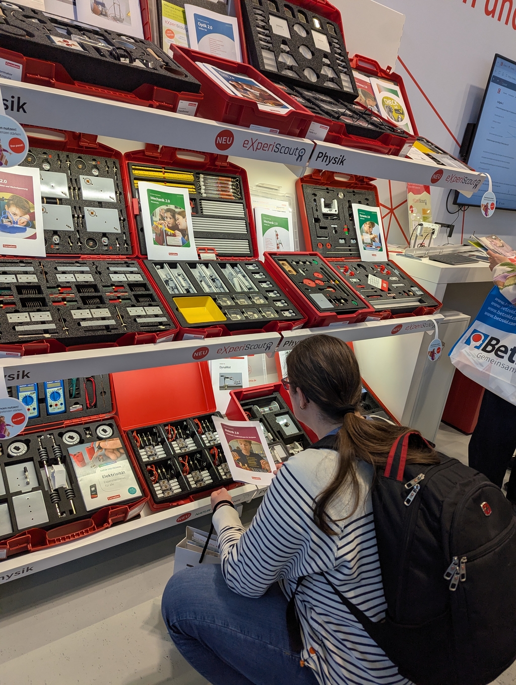 Eine Person betrachtet ein Regal mit Experimentierk&auml;sten und Zubeh&ouml;r f&uuml;r den Physikunterricht. Die farbenfrohen Koffer enthalten verschiedene Materialien und Instrumente, die f&uuml;r wissenschaftliche Experimente im Bildungsbereich genutzt werden k&ouml;nnen.