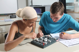 Zwei Schüler experimentieren mit einem elektrischen Schaltkreis, indem sie verschiedene Komponenten auf einer Tafel verbinden. Eine Person notiert ihre Beobachtungen auf einem Blatt Papier, während die andere konzentriert an den Verbindungen arbeitet.