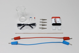 Ein Set aus physikalischem Experimentierbedarf liegt auf grauem Grund. Es umfasst eine Schutzbrille, ein Becherglas, eine Batterie, Klemmen, Bauteile sowie rote und blaue Anschlusskabel.